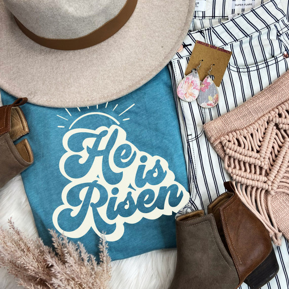 Cream He Is Risen Screen Print Heat Transfer, product image showing a blue cloth with a white 'He Is Risen' design, surrounded by fashion accessories including a beige hat, earrings, boots, a striped shirt, and a pink knitted blanket.