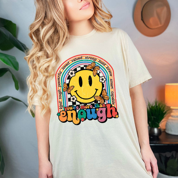 You're More Than Enough DTF Heat Transfer, colorful graphic design featuring a smiling yellow face, rainbow, butterflies, and inspirational words on a woman's t-shirt.