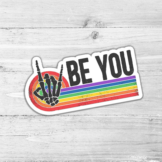 Be You Die Cut Sticker, colorful skeleton hand making a peace sign with rainbow stripes and the text 'BE YOU' on a white wooden background.