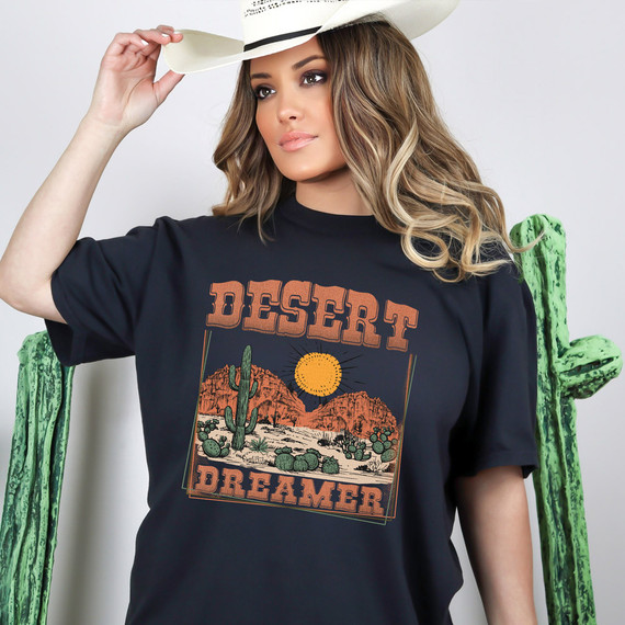 Desert Dreamer DTF Heat Transfer, desert landscape with cacti and mountains featuring a bright sun, on a woman's black t-shirt with wide-brimmed hat.