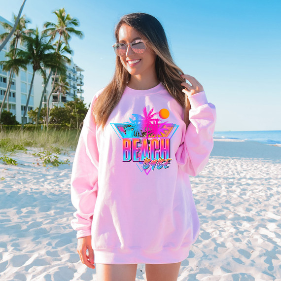 Beach Babe DTF Heat Transfer, a colorful beach-themed design with palm trees, the sun, and vibrant pink and blue colors on a pink sweatshirt worn by a woman on the beach.