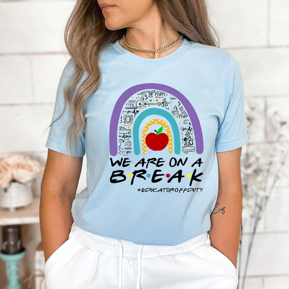 We Are On A Break #EducatorOffDuty Red Apple DTF Heat Transfer, woman wearing a light blue t-shirt with colorful rainbow and apple design, casual style with white pants and layered necklaces.