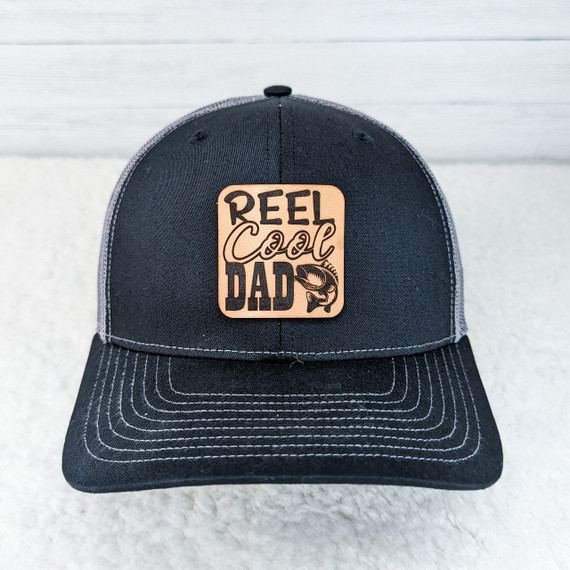Reel Cool Dad Leather Hat Patch, a patch with a wooden texture and the text 'Reel Cool Dad' along with a fish illustration, sewn onto a black and gray baseball cap.