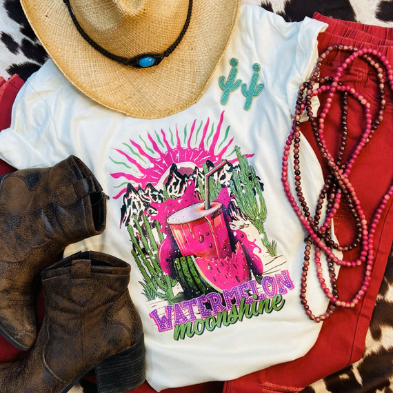 Watermelon Moonshine Screen Print Heat Transfer, colorful design featuring a watermelon drink with mountains and cactus, on a white T-shirt with accessories like a straw hat, beaded necklaces, boots, and red pants around.