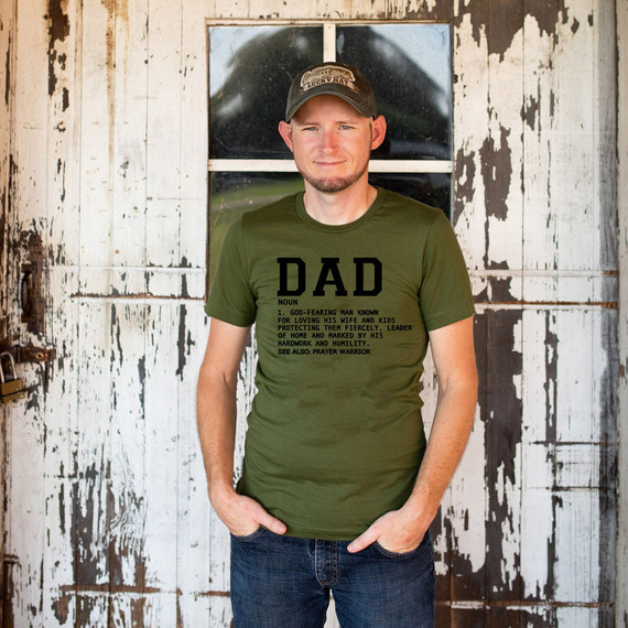 Dad Definition DTF Heat Transfer, a man wearing a green t-shirt with the word "DAD" and a definition printed on it, standing in front of a rustic wooden background.