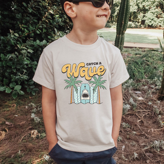 Catch A Wave DTF Heat Transfer, design featuring bold yellow text saying 'Catch A Wave' with palm trees and a cartoon shark in the ocean on a white t-shirt worn by a boy outdoors.