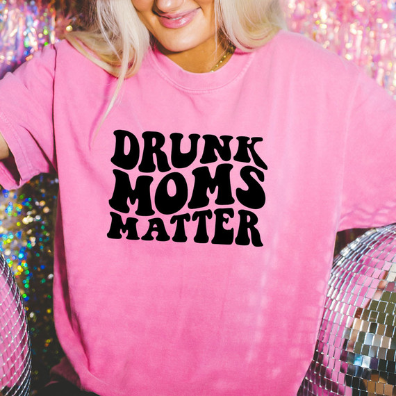 Drunk Moms Matter DTF Heat Transfer, close-up of a woman wearing a pink T-shirt with bold black text 'Drunk Moms Matter', standing against a colorful, sparkly background.