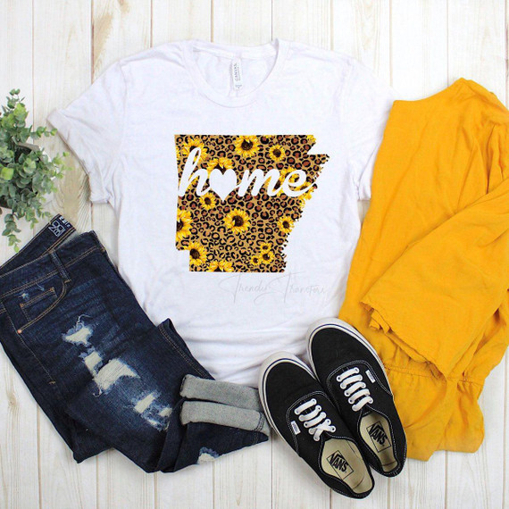 Arkansas Leopard and Sunflower with Home Sublimation Transfer T-shirt, white T-shirt featuring a graphic design of the state of Arkansas with sunflower and leopard print patterns, surrounded by denim jeans, black Vans shoes, yellow clothing, and a green plant on a light wooden background.