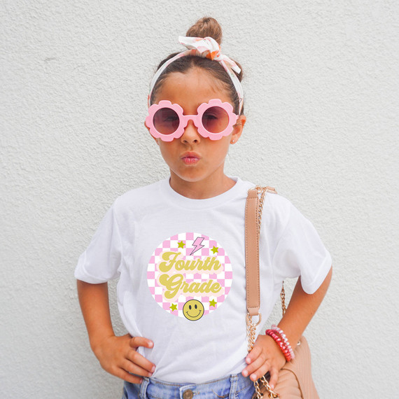 Fourth Grade Checkered DTF Heat Transfer, colorful design on a girl's white t-shirt featuring pink and white checkered background with yellow text, smiley face, stars, and lightning bolt.