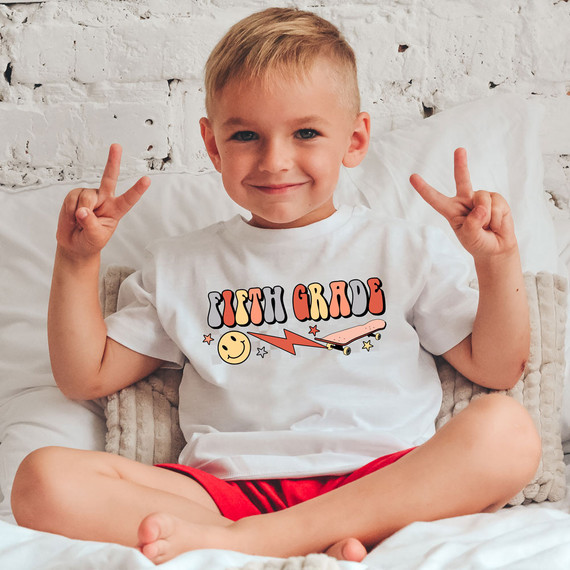 Fifth Grade Lightning Bolt DTF Heat Transfer, a colorful graphic design with the text 'Fifth Grade' along with a smiley face, lightning bolt, skateboard, and stars on a child's white T-shirt.