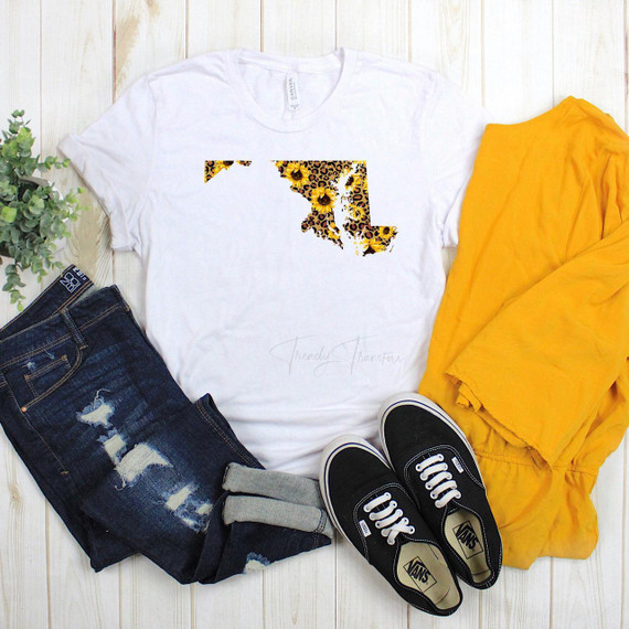 Maryland Leopard Sunflower Sublimation Transfer, casual outfit with white T-shirt featuring sunflower pattern in leopard print in the shape of Maryland state, ripped jeans, black Vans sneakers, yellow cloth, and a potted green plant on a wooden surface.