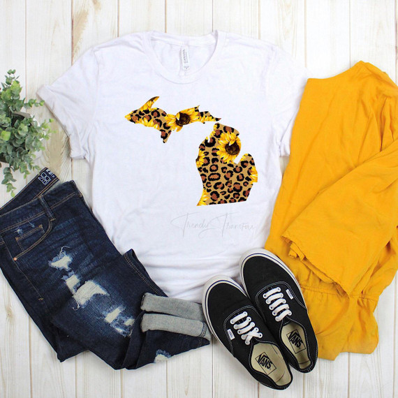 Michigan Leopard Sunflower Sublimation Transfer, white t-shirt with a leopard and sunflower pattern outline of Michigan, casual outfit with blue distressed jeans, black Vans sneakers, and a yellow jacket, on a light wooden surface.