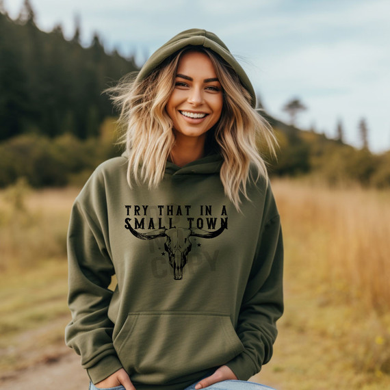 Try That In A Small Town Bullhead DTF Heat Transfer, a woman wearing an olive green hoodie with a bull skull graphic and the text 'Try That In A Small Town' printed on the front, outdoors in a natural setting.