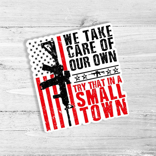 Try That In A Small Town Die Cut Sticker featuring a black rifle, stars, and stripes, with the text 'We Take Care Of Our Own' and 'Try That In A Small Town' in red and black, on a light wooden background.
