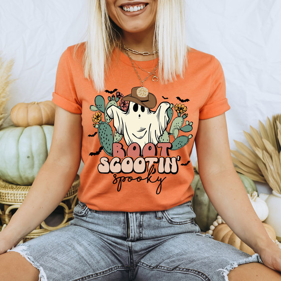 Boot Scootin' Spooky DTF Heat Transfer, Halloween-themed ghost with a cowboy hat surrounded by cacti and bats on an orange T-shirt.