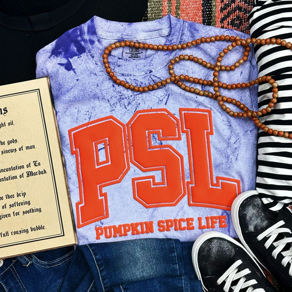 Pumpkin Spice Life Orange PUFF Screen Print Heat Transfer, close-up view of a purple tie-dye T-shirt with orange pumpkin spice life text and numbers, along with a string of beads, a book, sneakers, and striped clothing around it.