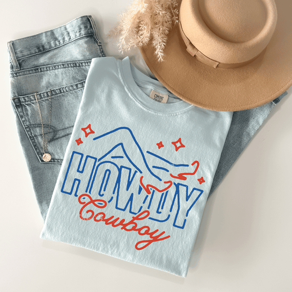 Howdy Cowboy DTF Heat Transfer, a graphic t-shirt with a cowboy hand-drawn design and bold text, paired with denim jeans and accessorized with a beige hat and dried flowers for stylish display.