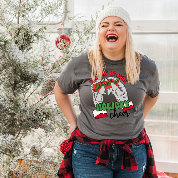 Rolling Up Some Holiday Cheer DTF Heat Transfer, a woman in a holiday-themed setting wearing a gray t-shirt with festive Christmas graphics and text, celebrating holiday cheer.