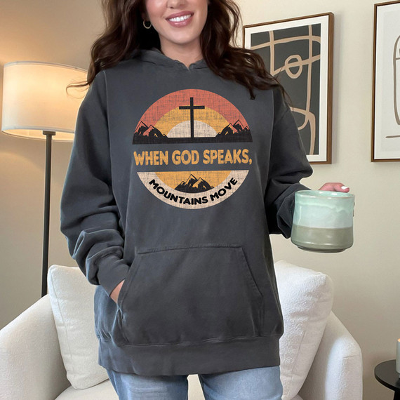 When God Speaks, Mountains Move DTF Heat Transfer, a woman wearing a hoodie with a mountain landscape and cross graphic design, with the slogan 'When God Speaks, Mountains Move' printed on it.