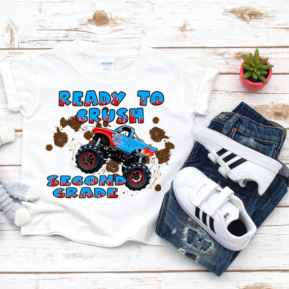 Back to School Monster Truck Crush the Second Grade Sublimation Transfer, white children's T-shirt with a colorful monster truck graphic and playful text, beside denim jeans, white sandals, and a small potted green plant on a white wooden surface.