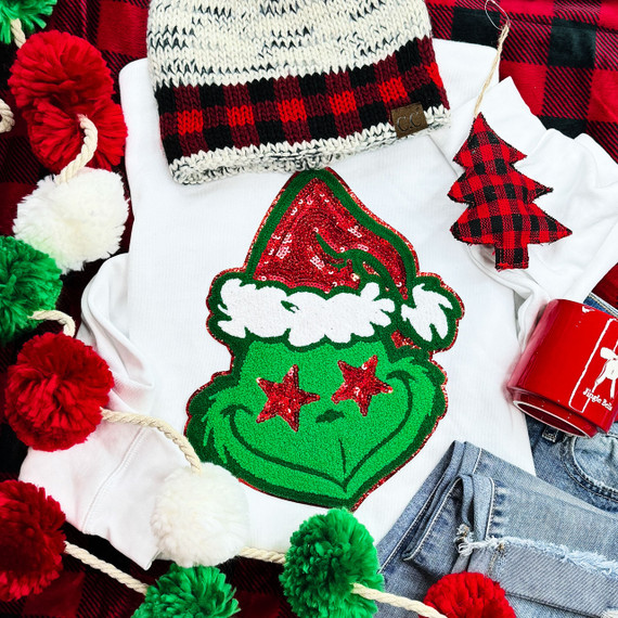 Grouchy Green Guy Chenille Patch, festive green Christmas elf face with red sequin hat and red star eyes on white fabric, surrounded by holiday decorations.