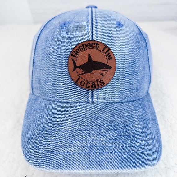 Respect The Locals Leather Hat Patch, showcasing a brown leather patch with a black shark silhouette and the text 'Respect The Locals' embroidered on a light blue denim baseball cap.