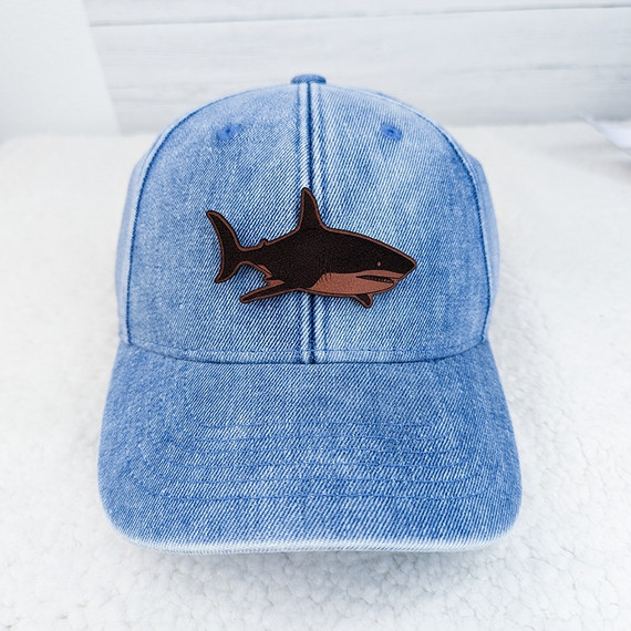 Shark Leather Hat Patch, a black and brown embroidered patch of a shark sewn onto a light blue denim baseball cap.