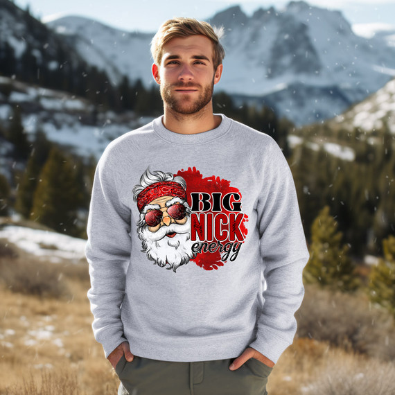 Big Nick Energy DTF Heat Transfer, a creative and vibrant graphic design featuring Santa Claus with sunglasses and a red bandana, placed on a light gray sweatshirt worn by a man outdoors with snow-capped mountains in the background.