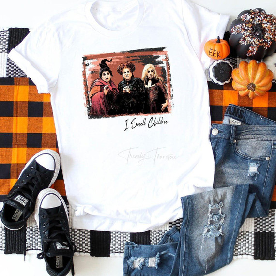 I Smell Children Sanderson Sisters Sublimation Transfer T-shirt, a white graphic tee featuring the Sanderson sisters from Hocus Pocus with the text I Smell Children, laid flat among Halloween decorations, pumpkins, and casual clothing.