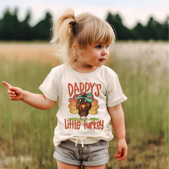Daddy's Little Turkey Bow DTF Heat Transfer, a cute design featuring a cartoon turkey with a bow on its head and the text 'Daddy's Little Turkey' on a child's t-shirt.