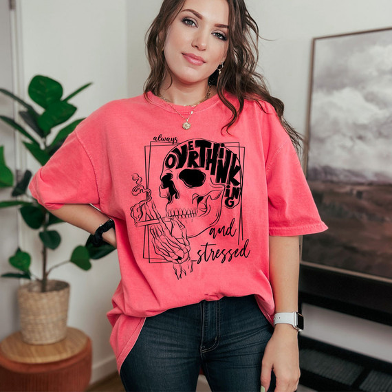 Always Overthinking And Stressed DTF Heat Transfer, a woman wearing a pink T-shirt with a skull and text design, standing indoors in a casual setting.