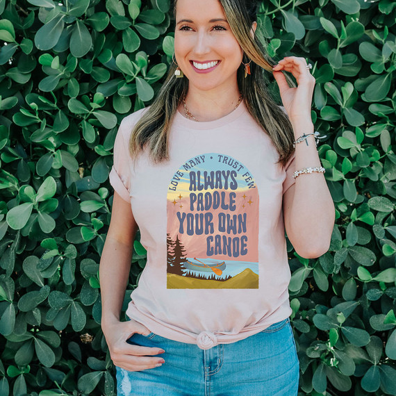 Always Paddle Your Own Canoe DTF Heat Transfer, a woman wearing a T-shirt with a colorful canoe and mountain design, standing in front of green foliage.