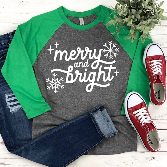 Merry And Bright WHITE DTF Heat Transfer, a white snowflake and festive lettering on a dark gray and green raglan shirt, with red sneakers and green foliage.