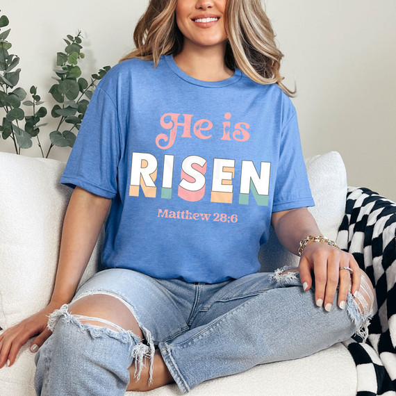 He Is Risen Matthew 28:6 DTF Heat Transfer, colorful and vibrant Bible verse design on a blue T-shirt worn by a woman sitting on a couch.