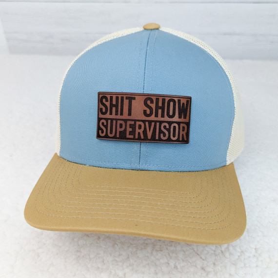 Shit Show Supervisor Leather Hat Patch, light blue and beige trucker hat with a rectangular brown and black embroidered patch that reads 'SHIT SHOW SUPERVISOR'.