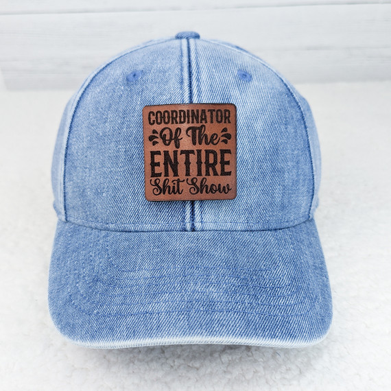 Coordinator Of The Entire Shit Show Leather Hat Patch, close-up image of a denim baseball cap with a brown leather patch that says "Coordinator Of The Entire Shit Show" in black lettering.