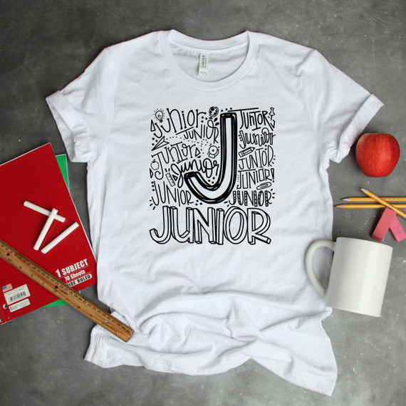 Junior Typography Word Art Sublimation Transfer, white t-shirt with black letter J designed with various lettering styles and small doodles, laid on a gray surface surrounded by school supplies and an apple.
