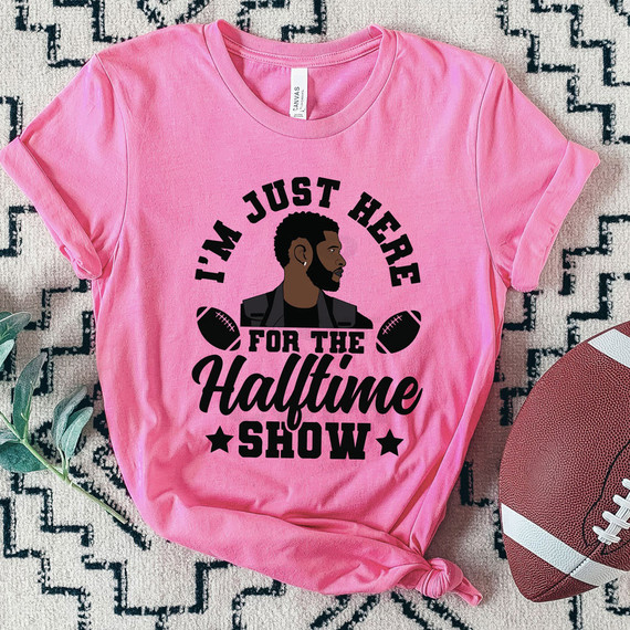I'm Just Here For The Halftime Show LVIII DTF Heat Transfer, pink T-shirt with a graphic of a man and footballs, and text about the halftime show.