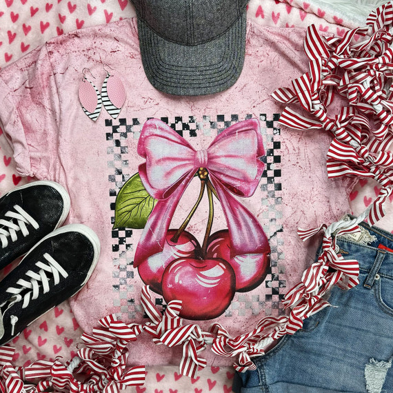 Cherries and Bow Screen Print Heat Transfer, pink graphic design of cherries with a large pink bow on a pink distressed T-shirt, surrounded by striped ribbon, striped earrings, black sneakers, a gray hat, and denim shorts.