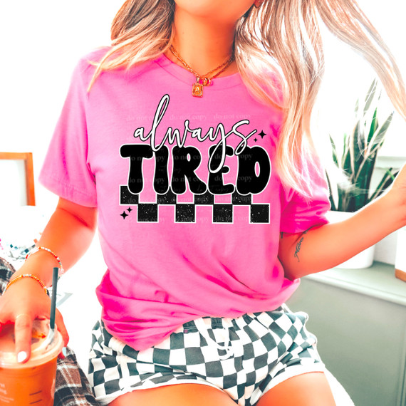 Always Tired DTF Heat Transfer, pink T-shirt with 'Always Tired' graphic design in black and white on the front, worn by a woman with blonde hair, tattoo, and jewelry, sitting indoors with a drink in hand.