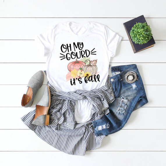 Oh My Gourd It's Fall Sublimation Transfer, a fall-themed white t-shirt with pumpkin and sunflower design and playful text, displayed alongside denim shorts, striped skirt, gray shoes, a plant, and a book on a white wooden background.