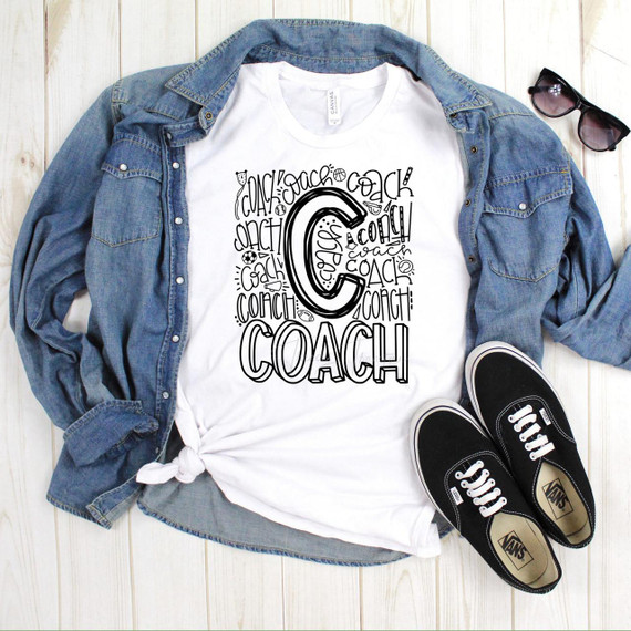 Coach Typography Sublimation Transfer, a white t-shirt with black hand-drawn style coach letter and logo design, laid flat with a denim jacket, black sunglasses, and black Vans sneakers on a wooden surface.