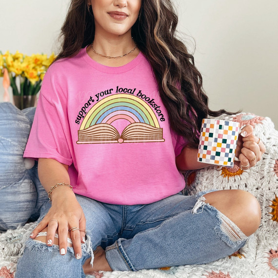 Support Your Local Bookstore DTF Heat Transfer, a colorful rainbow and open book design on a pink t-shirt worn by a woman sitting on a couch holding a patterned coffee mug.