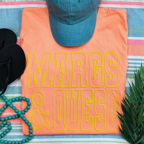 Margs & Queso Yellow PUFF Screen Print Heat Transfer, product flat lay with a yellow print design on a coral fabric, placed alongside accessories, a blue cap, a turquoise beaded necklace, and a pair of black flip flops on a colorful striped towel.