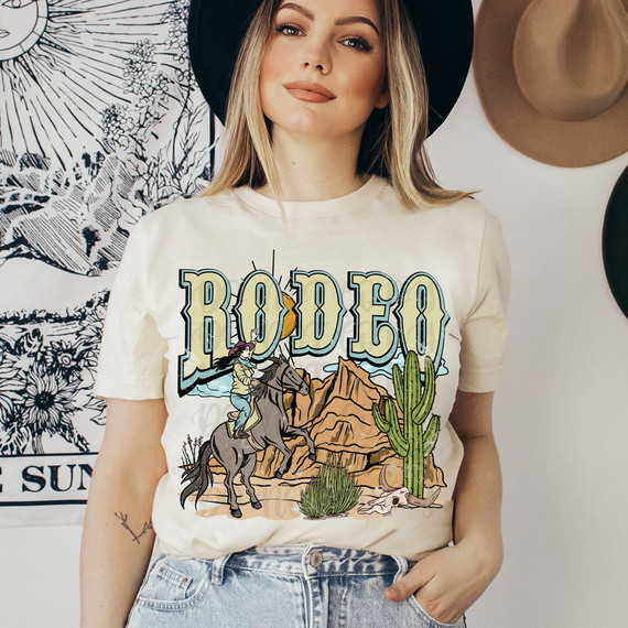 Rodeo Western DTF Heat Transfer, woman wearing a white T-shirt with rodeo-themed graphic, featuring a cowboy riding a horse, desert landscape, cacti, and mountains.