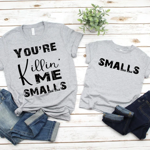 You're Killin' Me Smalls Mommy and Me Sublimation Transfer, matching grey T-shirts with funny text designs for mothers and children, paired with denim jeans on a white wooden surface.
