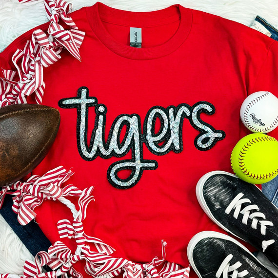 Tigers Sequin Patch, colorful sequin embroidery patch of a tiger for clothing decoration placed on a red sports shirt surrounded by sports accessories like baseballs, a pair of black sneakers, and a leather ball.