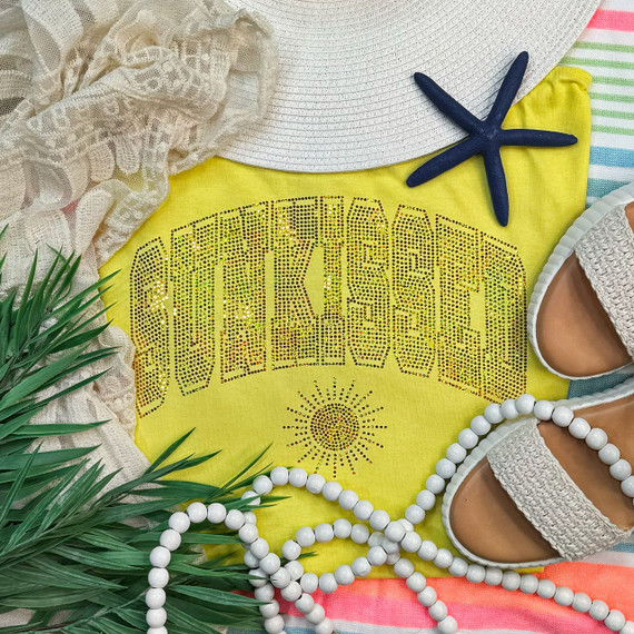 Sunkissed Spangled Transfer, a yellow fabric with rhinestone embellishments forming the word 'SUNKISSED' and a sun design, surrounded by beach accessories like a starfish, hat, sandals, a string of white beads, and a lace fabric, creating a summer beach scene.
