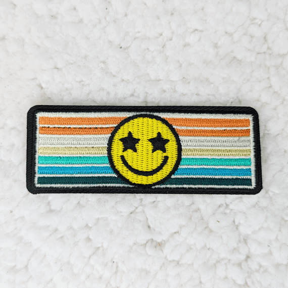 Retro Happy Face Embroidered HAT/POCKET Patch, colorful embroidered patch with a yellow smiley face having star-shaped eyes on a striped rainbow background.