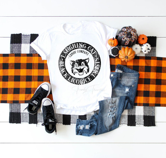 Laughing cat candy company Sublimation Transfer, a printed design of a laughing cat logo on a white T-shirt, displayed alongside Halloween-themed pumpkins, a pair of distressed jeans, a checkerboard and plaid table runner, and black sneakers.